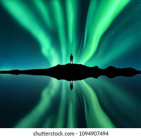 オーロラと立っている男のシルエット。ロフォーテン諸島、ノルウェー。オーロラと幸せな男。星と緑の極光。オーロラ、男、湖、水の反射と夜の風景。トラベル