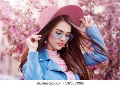Joven hermosa chica de moda con elegantes gafas de sol de aviador de color azul, sombrero de gamuza rosa, aretes, chaqueta de motorista. Modelo posando en la calle con árboles en flor. Concepto de moda de primavera.