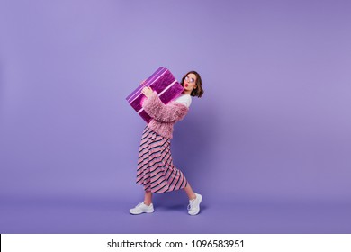 Mujer joven adorable en zapatillas blancas posando con caja de regalo. Retrato de estudio de una chica fascinante haciendo cara de besos expresando mientras sostiene un gran regalo.