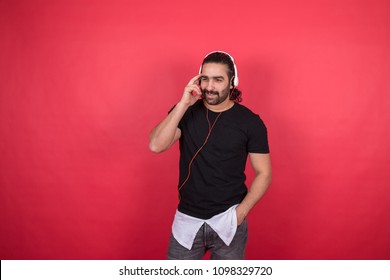 Knappe man draagt ​​een casual outfit, luistert naar muziek, staat en legt een hand op zijn zak en de andere op zijn voorhoofd, staande op een rode achtergrond.