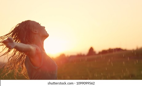 NAHAUFNAHME, LENS FLARE, COPY SPACE: Eine lächelnde junge Frau dreht sich mit ausgestreckten Armen im Regen. Das blonde kaukasische Mädchen tanzt spielerisch bei atemberaubendem Sonnenuntergang. Tänzerin improvisiert im Regen