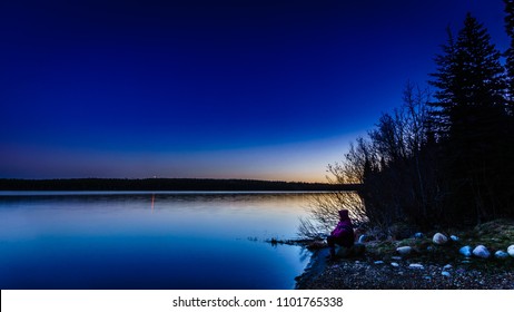 Letztes Licht über einem friedlichen See, während der Tag in die Nacht übergeht Einsame Person, die am Seeufer sitzt.