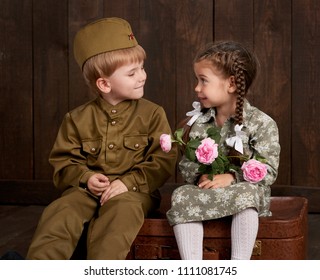 djeca dječak su obučeni kao vojnici u retro vojnim uniformama i djevojčica u ružičastoj haljini sjedi na starom koferu, tamna drvena pozadina, retro stil