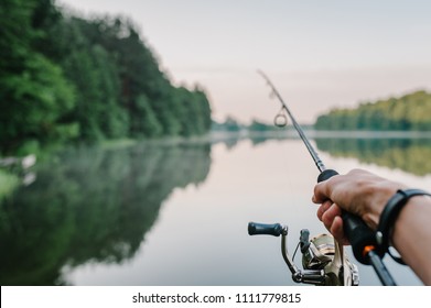 川岸で釣り竿、スピニング リールを持つ漁師。日の出。カワカマス、スズキ、コイの釣り。湖を背景に霧。背景霧の朝。野生の自然。田舎の休暇の概念。