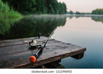 背景の桟橋の川岸に釣り竿、スピニング リール。日の出。湖を背景に霧。霧の深い朝。野生の自然。田舎の休暇の概念。釣りの日についての記事。