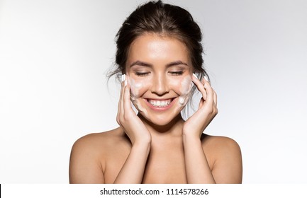 Retrato de una alegre mujer que se ríe aplicando espuma para lavarse la cara. Hermosa morena con apariencia atractiva. Concepto de relajación del spa para el cuidado de la piel. aislado sobre fondo gris