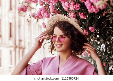 Retrato al aire libre de una joven hermosa y feliz sonriente chica rizada con elegantes gafas de sol rosas, sombrero de paja, vestido. Modelo posando en la calle cerca de rosas florecientes. Concepto de moda de verano