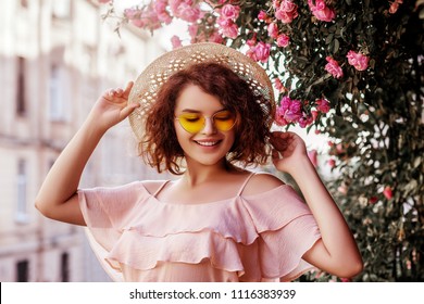 Outdoor close up portrait of young beautiful happy smiling curly girl wearing stylish yellow sunglasses, straw hat, pink blouse with ruffles. Model posing near blooming roses. Summer fashion concept
