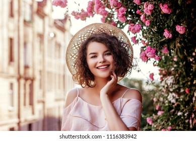 Retrato al aire libre de una joven hermosa y feliz sonriente chica rizada con elegante sombrero de paja, blusa rosa con volantes. Modelo posando en la calle cerca de rosas florecientes. Concepto de moda de verano. copia espacio