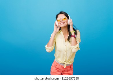 Modelo asiática inspirada con reloj de pulsera rosa y pulsera verde escuchando música sobre fondo azul. Fotografía en interiores de una chica latina extasiada con gafas de sol naranjas tocando auriculares y sonriendo.