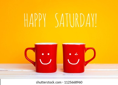 Two red coffee mugs with a smiling faces on a yellow background with phrase Happy saturday. Happy coffee mugs.