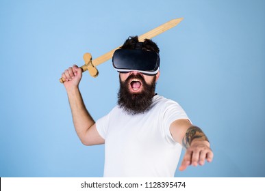 Hipster trên khuôn mặt hét lên thích chơi trò chơi trong thực tế ảo. Khái niệm game thủ VR. Chàng trai với màn hình gắn đầu và trò chơi chiến đấu chơi kiếm trong VR. Người đàn ông có râu trong kính VR, nền xanh nhạt.