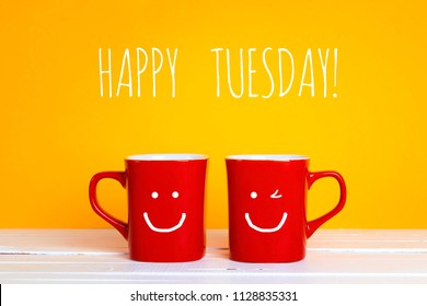 Two red coffee mugs with a smiling faces on a yellow background with phrase Happy tuesday. Happy coffee mugs.
