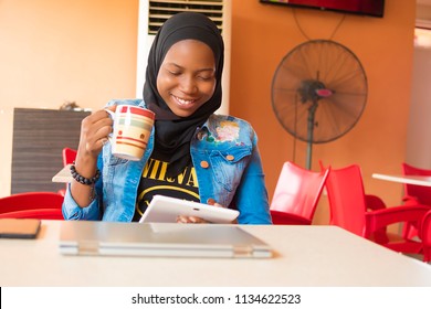 junge nigerianische muslimische Frau, die allein in einem Café sitzt und lächelt, während sie ihre Android-Tablette benutzt und eine Kaffeetasse hält