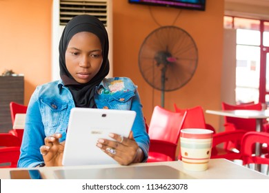 junge nigerianische muslimische Frau, die allein in einem Café sitzt und lächelt, während sie ihre Android-Tablette benutzt