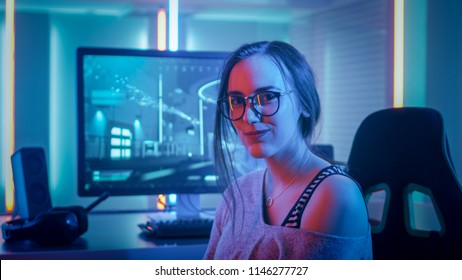 Porträt des schönen jungen Pro-Gamer-Mädchens, das an ihrem PC sitzt und in die Kamera schaut. Attraktive Geek Girl Spielerin mit Brille im Raum von Neonlichtern beleuchtet.