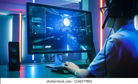Back View Shot of Professional Gamer Playing in First-Person Shooter Online Video Game on His Personal Computer. He's Talking with His Team Through Headset. Room Lit by Neon Lights in Retro Style.