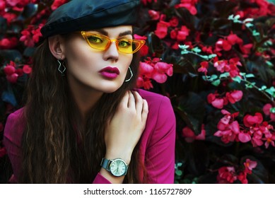 Nahaufnahme-Modeporträt der jungen schönen Frau im Freien, die trendige gelbe Sonnenbrille, Leder-Baskenmütze, Armbanduhr, Fuchsia-Farbblazer trägt und nahe blühenden rosa Blumen aufwirft. Kopieren, leerer Raum