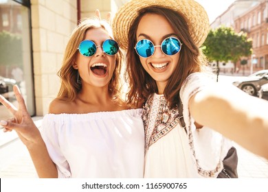 通りの背景にスマート フォンでソーシャル メディアの selfie 写真を撮る夏の白い流行に敏感なドレスで 2 つの若い女性笑顔ヒッピー ブルネットとブロンドの女性モデル。驚きの顔、感情、