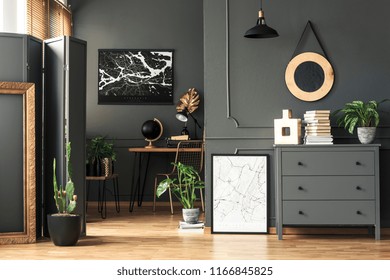 Black map on grey wall in dark living room interior with plants and poster. Real photo