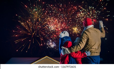 Happy New Year Celebration, Young Family of Three Standing in the Front Yard Watching Beautiful Fireworks. In the Evening while Snow is Falling Father, Mother and Cute Little Daughter Look up.