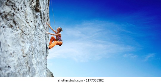 Seitenansicht der jungen schlanken Klettererin in leuchtend orangefarbenen Hosen, die auf der Klippe klettern. Eine Frau klettert auf eine vertikale Felswand auf dem Hintergrund des blauen Himmels
