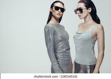 Dvě ženy nosí stříbrné módní oblečení se slunečními brýlemi na šedém pozadí. Modelka ve futuristickém vzhledu ve studiu.