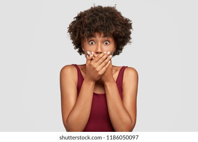 Shocked Afro American woman tries be speechless and mute, keeps hands on mouth, recieves bad news from interlocutor, wears casual outfit, stares with bugged eyes at camera, isolated over white wall