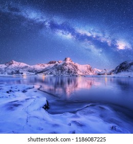 Milchstraße über gefrorener Meeresküste und schneebedeckte Berge im Winter nachts auf den Lofoten-Inseln, Norwegen. Arktische Landschaft mit blauem Sternenhimmel, Wasser, Eis, schneebedeckten Felsen, Milchstraße. Schöner Raum