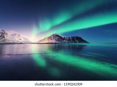 Nordlichter auf den Lofoten, Norwegen. Grüne Nordlichter. Sternenhimmel mit Polarlichtern. Nachtwinterlandschaft mit Aurora, Meer mit Himmelsreflexion, Felsen, Strand und schneebedeckten Bergen. Natur
