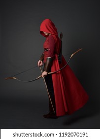 Porträt des brünetten Mädchens in voller Länge, das rotes mittelalterliches Kostüm und Umhang trägt und Pfeil und Bogen hält. stehende Pose auf grauem Studiohintergrund.
