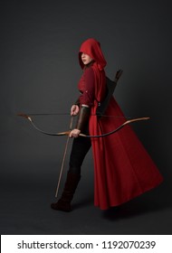 Porträt des brünetten Mädchens in voller Länge, das rotes mittelalterliches Kostüm und Umhang trägt und Pfeil und Bogen hält. stehende Pose auf grauem Studiohintergrund.