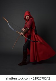 Porträt des brünetten Mädchens in voller Länge, das rotes mittelalterliches Kostüm und Umhang trägt und Pfeil und Bogen hält. stehende Pose auf grauem Studiohintergrund.