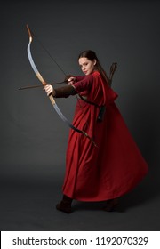 Porträt des brünetten Mädchens in voller Länge, das rotes mittelalterliches Kostüm und Umhang trägt und Pfeil und Bogen hält. stehende Pose auf grauem Studiohintergrund.