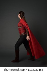 full length portrait of brunette girl wearing red medieval costume and cloak. standing pose with back to the camera on grey studio background.