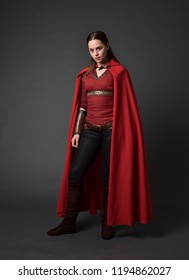 full length portrait of brunette girl wearing red medieval costume and cloak. standing pose on grey studio background.