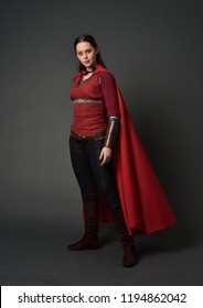 full length portrait of brunette girl wearing red medieval costume and cloak. standing pose on grey studio background.