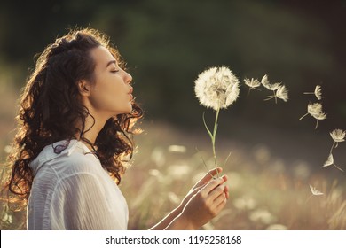 Wanita Muda Cantik duduk di lapangan di rumput hijau dan meniup dandelion. Di luar rumah. Menikmati alam. Gadis Tersenyum Sehat di halaman musim panas. Konsep bebas alergi. Ras campuran langsing cantik bule