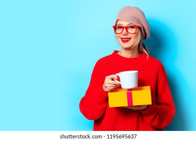 Portrait of a young girl in red sweater with gift box and cup of coffee on blue background