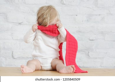 Binnenportret van een schattige blonde baby die een rode wollen sjaal draagt, een kiekeboe-spel speelt of geen concept wil aankleden