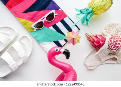Plat leggen van zomerse vibes concept met kleurrijke reismodeartikelen, zonnebril, sjaal, roze drakenfruit, flamingo opblaasbare bekerhouder, ananasfles, nettas en sandaal op een witte achtergrond