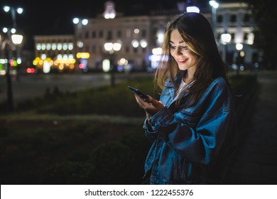 夜の街で携帯電話にテキスト メッセージを送信する若い白人女性