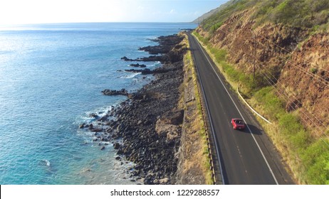 AERIAL: Red convertible car driving along the picturesque coastal road above the great ocean cliffs and sea water splashing into rocks. Happy young couple on summer vacation traveling at the seaside
