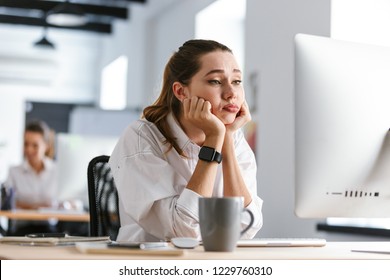 Mujer joven aburrida vestida con camisa sentada en su lugar de trabajo en la oficina, mirando la computadora