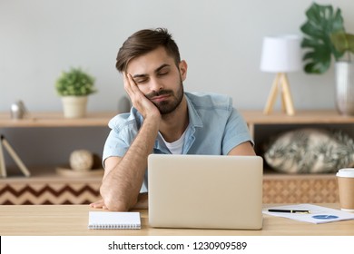 Un hombre milenario exhausto sostiene la cabeza con la mano sentado en la mesa de la oficina quedándose dormido, un hombre cansado se queda dormido trabajando en una laptop en el lugar de trabajo de su casa, un tipo somnoliento siente fatiga tomando una siesta cerca de la computadora