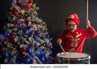 カーニバルの衣装を着た幸せな白人の幼稚なドラマー、サンタクロースの鹿が、ドラムスティックを手にした新しいドラムセットで演奏します。両親はクリスマスの贈り物として子供の太鼓を買った. クリスマスツリーのライト