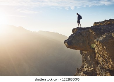 日の出の崖の端に立っているバックパックを持つ若い観光客。オマーンのグランドキャニオン、ジェベル・アクダル。