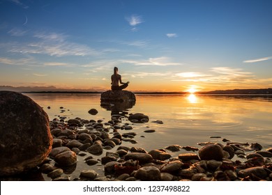 young beautiful woman girl are doing sport fitness yoga position on a stone at a beautiful lake, sea, see in the water - in the background you can see a colorful sundown - stone, water, orange sundown