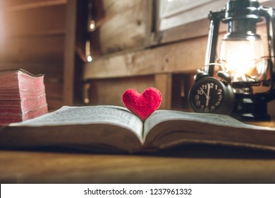Otvorená Biblia a červené srdce v strede so svetlom olejovej lampy doma. ráno. na drevenom stole. pojem pre lásku k Bohu.