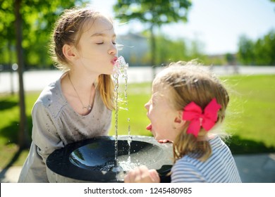 Two sisters having fun with drinking water fountain on warm and sunny summer day. Kids playing outdoors.